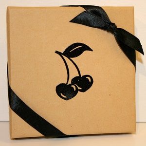 Peut inclure: Une bo&icirc;te cadeau en papier brun avec un ruban et un n&oelig;ud noirs. La bo&icirc;te a une silhouette noire de deux cerises avec une tige et une feuille sur le devant.