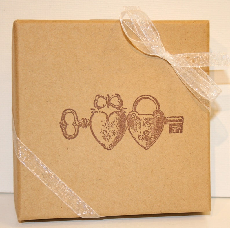 May include: A brown cardboard box with a white ribbon tied around it. The box features a vintage-style illustration of a key, a padlock, and two hearts.