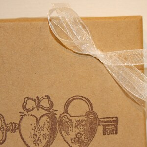 May include: A brown paper gift box with a white ribbon tied in a bow. The box features a vintage design with a key, a heart, and a padlock printed on the front.