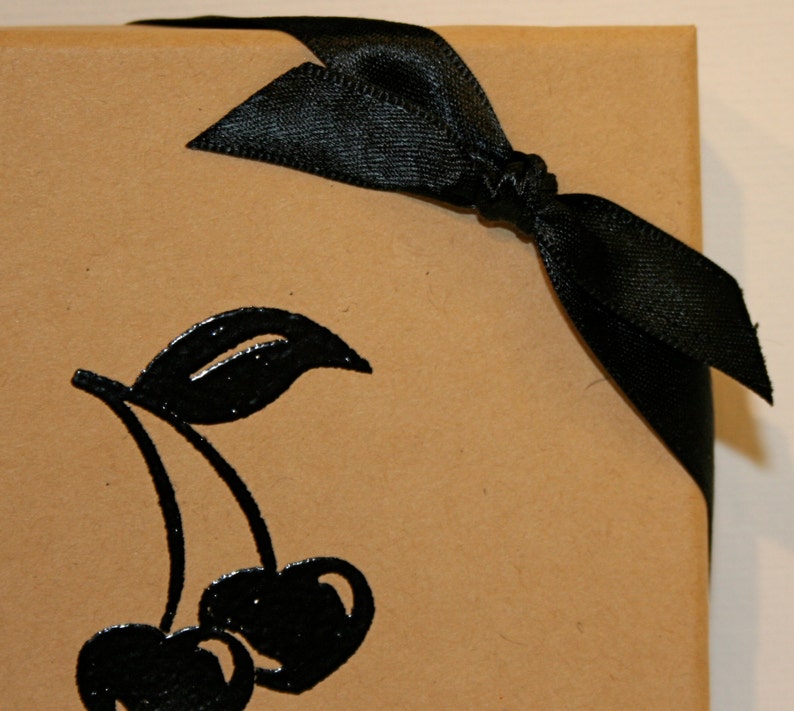 Peut inclure: Un sac cadeau en papier brun avec un ruban de satin noir nou&eacute; en un n&oelig;ud. Le sac a une silhouette noire de deux cerises avec une tige et une feuille.