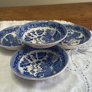 May include: Four small, round, blue and white ceramic bowls with a detailed, traditional pattern. The bowls are arranged on a white embroidered cloth, with a wooden surface in the background. The bowls have a classic, vintage aesthetic.