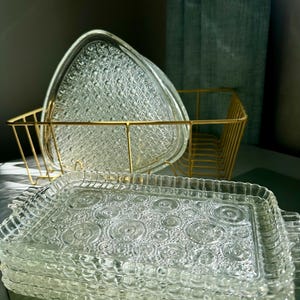 May include: A stack of clear glass serving trays with a textured, circular pattern. A gold wire basket holds triangular glass plates with a starburst design. The trays and plates are stacked, suggesting a set for serving food or displaying items.