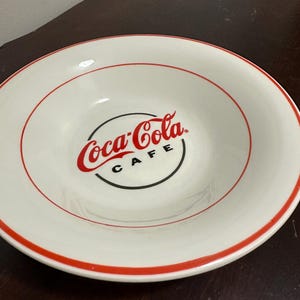 May include: White ceramic bowl with a red rim and the Coca-Cola Cafe logo in the center. The logo features the Coca-Cola script in red, with "CAFE" in black below. The bowl has a simple, classic design.