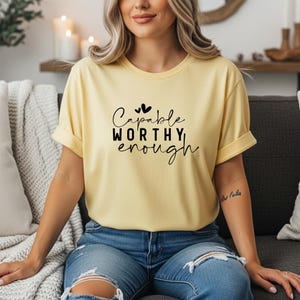 May include: A light yellow t-shirt with rolled-up sleeves. The shirt features the words "Capable, Worthy, Enough" in a black script font with two small heart graphics above the word "Capable". The person is wearing blue jeans.