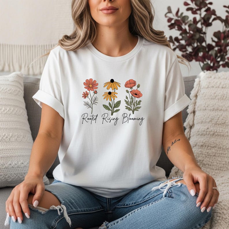 May include: White t-shirt featuring a floral design with three different flower illustrations in shades of orange, yellow, and red. The text "Rooted Rising Blooming" is printed below the flowers in a black script font.