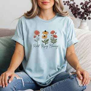 May include: Light blue t-shirt featuring a floral design with orange, yellow, and red flowers. The text "Rooted Rising Blooming" is printed below the flowers. The shirt is made of a soft material and has rolled-up sleeves.