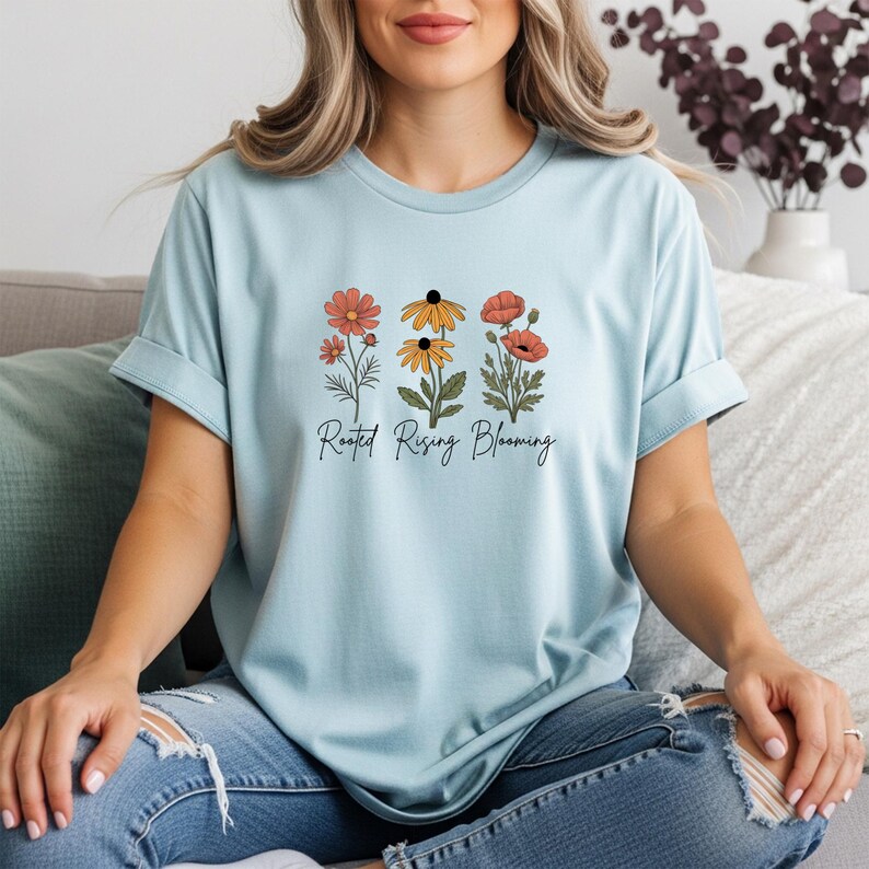 May include: Light blue t-shirt featuring a floral design with orange, yellow, and red flowers. The text "Rooted Rising Blooming" is printed below the flowers. The shirt is made of a soft material and has rolled-up sleeves.