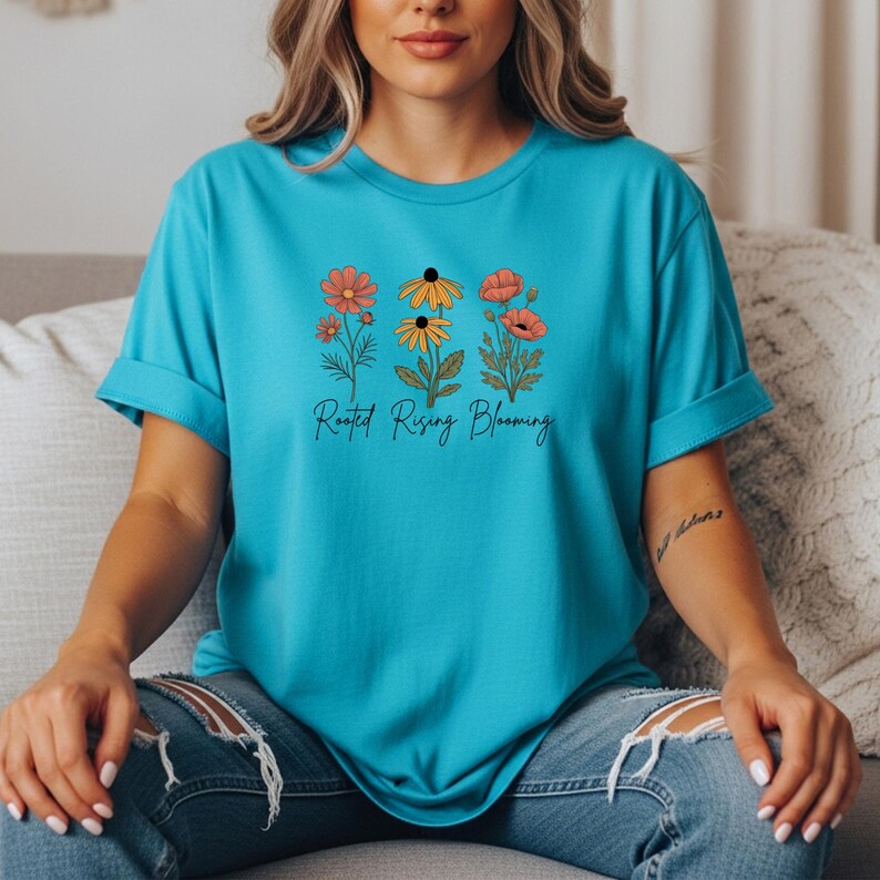 May include: A turquoise t-shirt featuring a floral design with orange, yellow, and red flowers. The words "Rooted Rising Blooming" are printed below the flowers in a script font. The shirt has rolled-up sleeves.