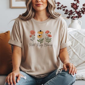 May include: Beige t-shirt featuring a floral design with three different flower types in shades of red, yellow, and orange. The text "Rooted Rising Blooming" is printed below the flowers in a script font. The shirt has short sleeves.