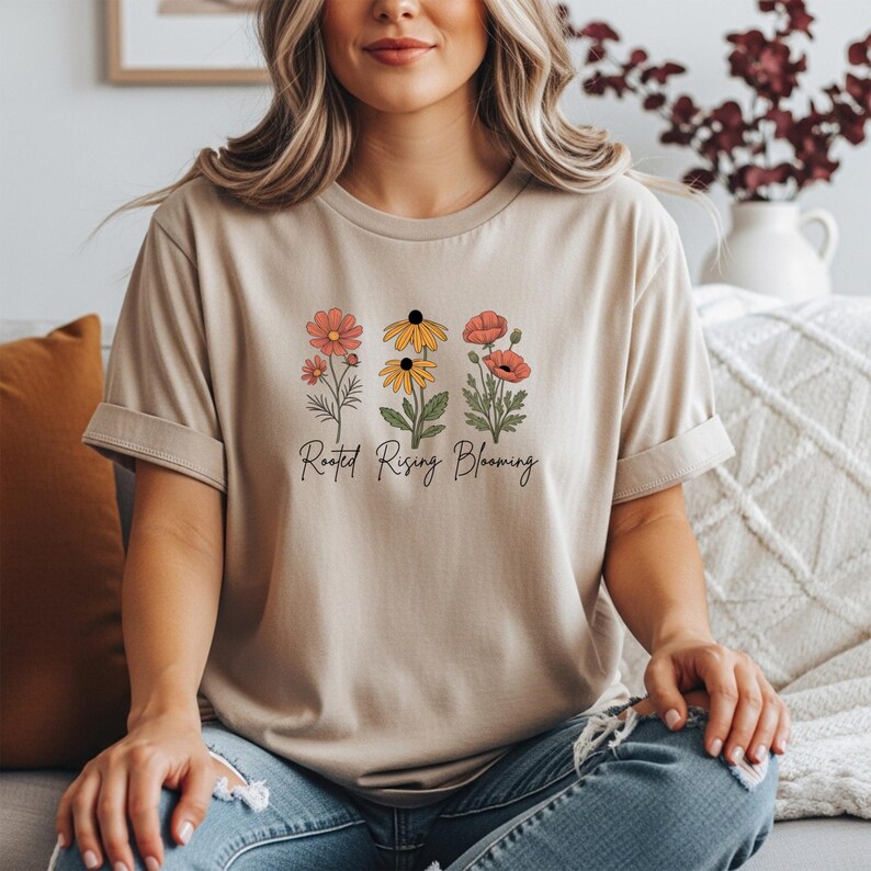 May include: Beige t-shirt featuring a floral design with three different flower types in shades of red, yellow, and orange. The text "Rooted Rising Blooming" is printed below the flowers in a script font. The shirt has short sleeves.