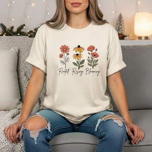May include: Cream-colored t-shirt featuring a floral design with three different flower illustrations in shades of red, yellow, and orange. The text "Rooted Rising Blooming" is printed below the flowers in a script font. The shirt is made of a soft material.