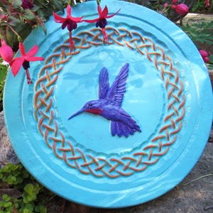 May include: A round, turquoise bird bath with a raised hummingbird design in the center. The hummingbird is purple with red accents. An orange Celtic knot design borders the bird bath. Fuchsia flowers are in the background.