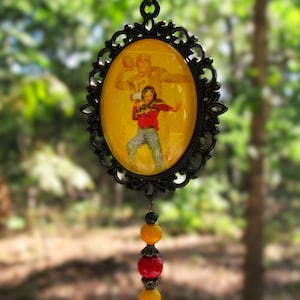 May include: An oval pendant featuring a black filigree frame and a yellow insert depicting a football player in a red jersey and blue jeans. Below the pendant hangs a vertical strand of three beads in alternating yellow and red colors.