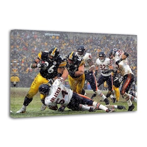 May include: A canvas print of an American football game in progress. Players in black and gold uniforms are running with the ball, while others in white and orange uniforms attempt to tackle them. The background is filled with spectators.