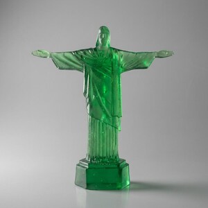 May include: A translucent green statue of Christ the Redeemer with outstretched arms. The statue is mounted on a square base and stands against a neutral background. The sculpture's details are visible, including the folds of the robes.