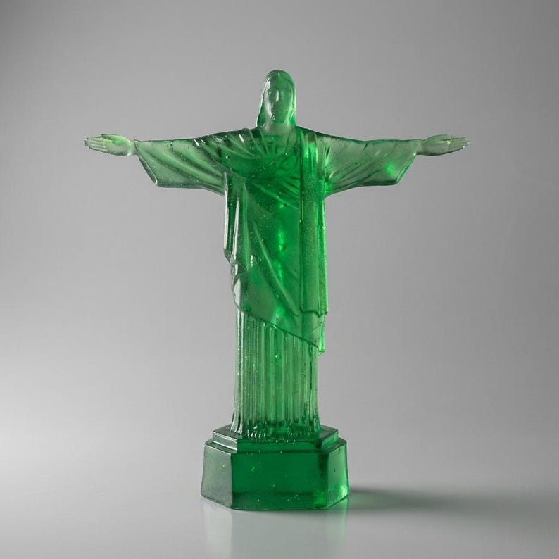May include: A translucent green statue of Christ the Redeemer with outstretched arms. The statue is mounted on a square base and stands against a neutral background. The sculpture's details are visible, including the folds of the robes.