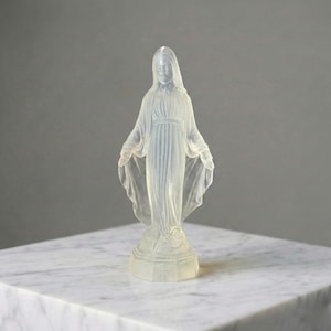 May include: A translucent, frosted glass figurine of a standing figure with a veiled head, wearing a long, flowing robe with gathered folds. The figure has arms extended outward with palms facing forward, standing on a circular base.