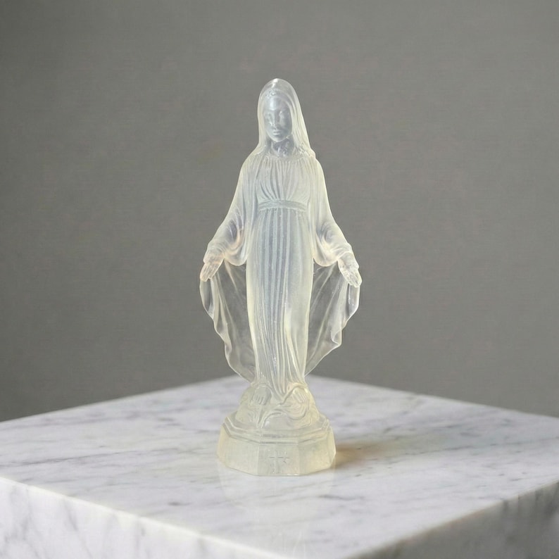 May include: A translucent, frosted glass figurine of a standing figure with a veiled head, wearing a long, flowing robe with gathered folds. The figure has arms extended outward with palms facing forward, standing on a circular base.