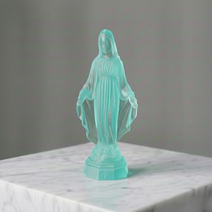 May include: A translucent, light blue frosted glass figurine depicting a standing figure with arms outstretched, wearing a long, draped robe. The figure stands on a hexagonal base featuring a small, embossed cross detail on the front panel.