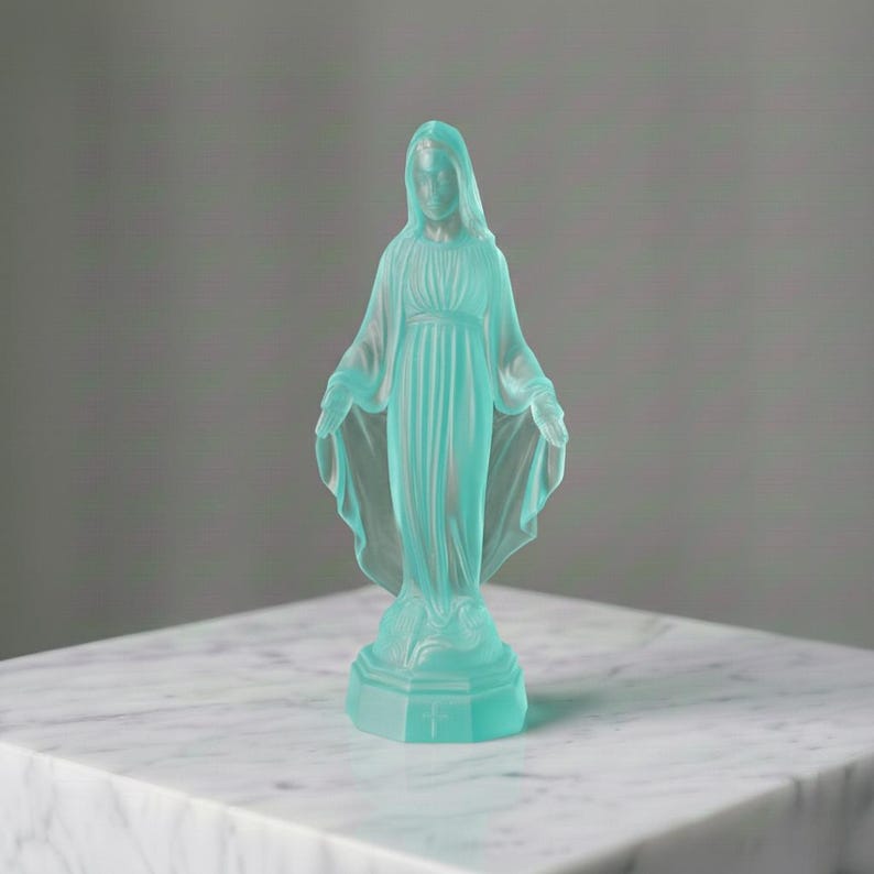 May include: A translucent, light blue frosted glass figurine depicting a standing figure with arms outstretched, wearing a long, draped robe. The figure stands on a hexagonal base featuring a small, embossed cross detail on the front panel.