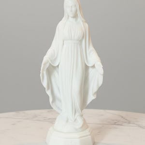 May include: A white statue of the Virgin Mary standing on a hexagonal base. The figure wears a long, flowing robe with draped folds and a head covering. The arms are extended outward with palms facing forward, and the feet rest upon a small mound.