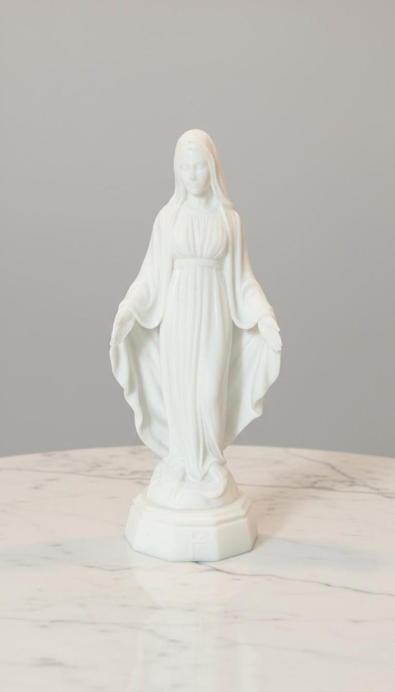 May include: A white statue of the Virgin Mary standing on a hexagonal base. The figure wears a long, flowing robe with draped folds and a head covering. The arms are extended outward with palms facing forward, and the feet rest upon a small mound.