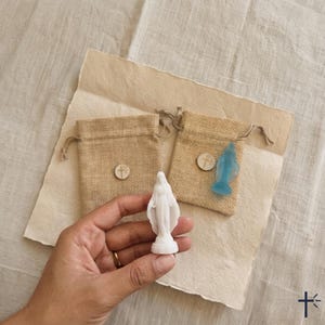 May include: A small white figurine of the Virgin Mary held in a hand, accompanied by two tan burlap drawstring pouches. Each pouch features a small circular wooden charm with a cross symbol. A translucent blue figurine is visible on one pouch.