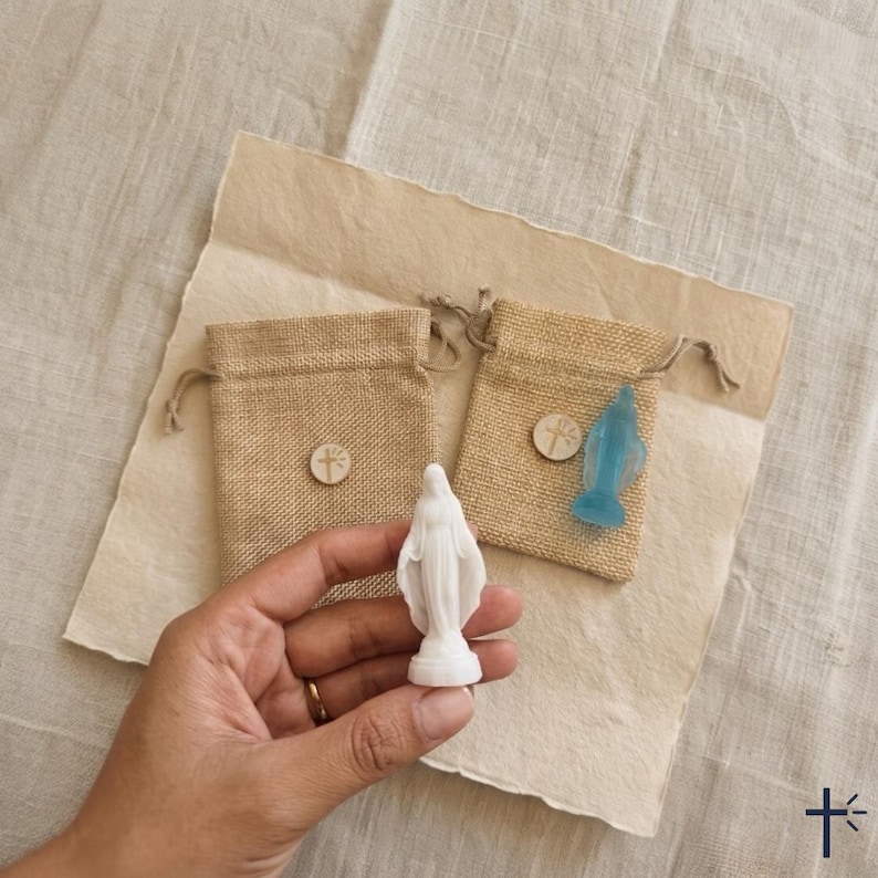 May include: A small white figurine of the Virgin Mary held in a hand, accompanied by two tan burlap drawstring pouches. Each pouch features a small circular wooden charm with a cross symbol. A translucent blue figurine is visible on one pouch.