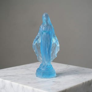 May include: A translucent light blue glass figurine of a standing figure with a draped head covering and long robes. The figure stands on a hexagonal base with a small cross symbol embossed on the front, featuring a smooth, frosted finish.