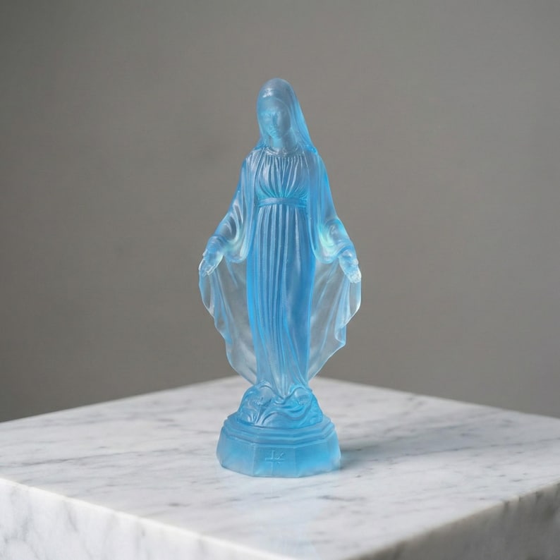 May include: A translucent light blue glass figurine of a standing figure with a draped head covering and long robes. The figure stands on a hexagonal base with a small cross symbol embossed on the front, featuring a smooth, frosted finish.