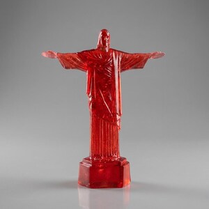 May include: A translucent red statue of Christ the Redeemer with outstretched arms. The statue is mounted on a rectangular base and stands against a gray background. The statue is a detailed replica of the iconic landmark.