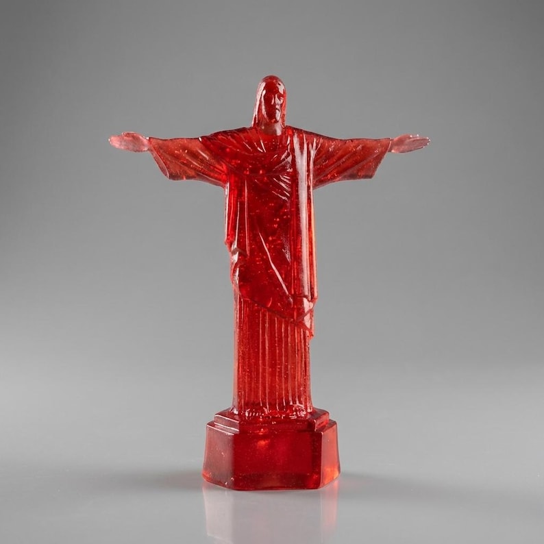 May include: A translucent red statue of Christ the Redeemer with outstretched arms. The statue is mounted on a rectangular base and stands against a gray background. The statue is a detailed replica of the iconic landmark.