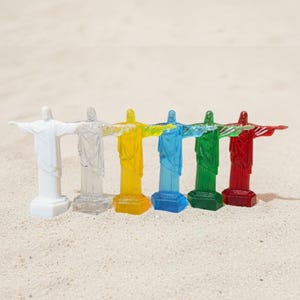 May include: Six miniature statues of Christ the Redeemer in a row, each in a different color: white, clear, yellow, blue, green, and red. The statues are standing on a sandy surface.