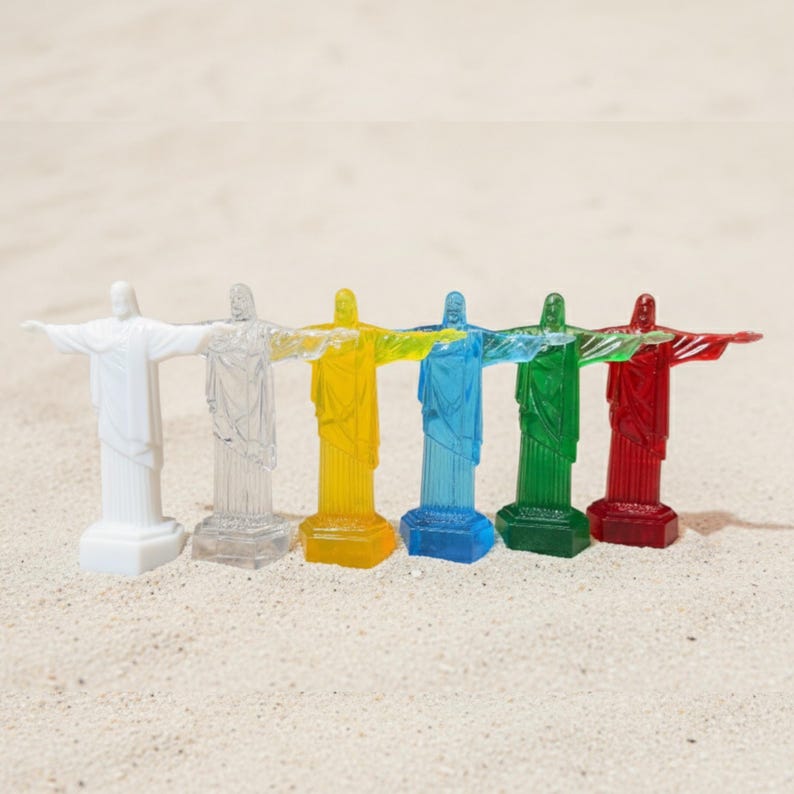 May include: Six miniature statues of Christ the Redeemer in a row, each in a different color: white, clear, yellow, blue, green, and red. The statues are standing on a sandy surface.