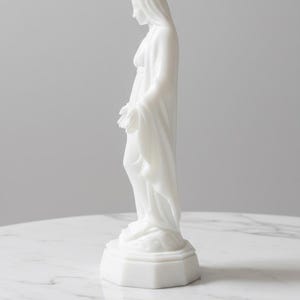 May include: A white, opaque, smooth-surfaced figurine of a standing figure with a draped head covering and long robes. The figure is positioned in profile, facing downward with hands slightly extended, resting on a hexagonal base.