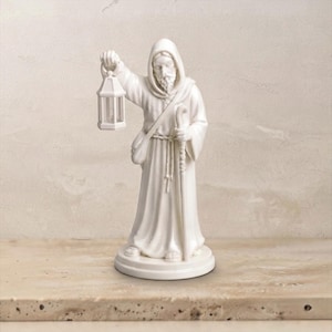 Könnte beinhalten: Eine weiße Keramikfigur eines bärtigen Mannes in einer Kapuzenrobe mit Sandalen. Die Figur hält eine Laterne in der rechten Hand und einen langen Wanderstab in der linken Hand. Eine Umhängetasche ist über die Brust gelegt und ein Seilgürtel um die Taille.