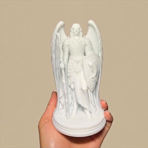 May include: A white figurine of an angel standing on a circular base. The figure wears detailed armour, possesses large feathered wings, and holds a tall staff in the right hand and a shield featuring a serpent emblem in the left hand.