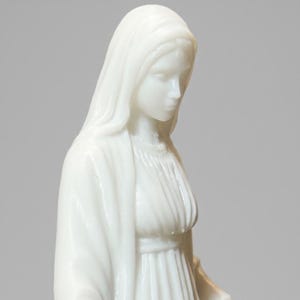 May include: A white figurine depicting a figure with a draped head covering and a long, pleated gown. The sculpture features smooth, glossy surfaces and soft, rounded facial features, capturing a serene expression with eyes cast downward.