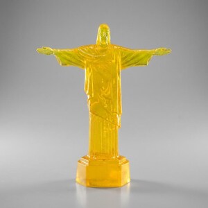 May include: A translucent yellow statue of Christ the Redeemer with outstretched arms. The statue is on a rectangular base and stands on a gray surface. The statue is a miniature replica of the famous landmark.
