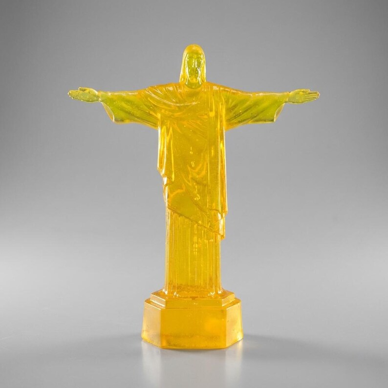 May include: A translucent yellow statue of Christ the Redeemer with outstretched arms. The statue is on a rectangular base and stands on a gray surface. The statue is a miniature replica of the famous landmark.