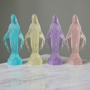 May include: Four translucent statues of the Virgin Mary standing in a row, each featuring a different pastel color: cyan, pale yellow, lavender, and soft pink. Each figure is depicted with arms outstretched, wearing a draped robe on a base.