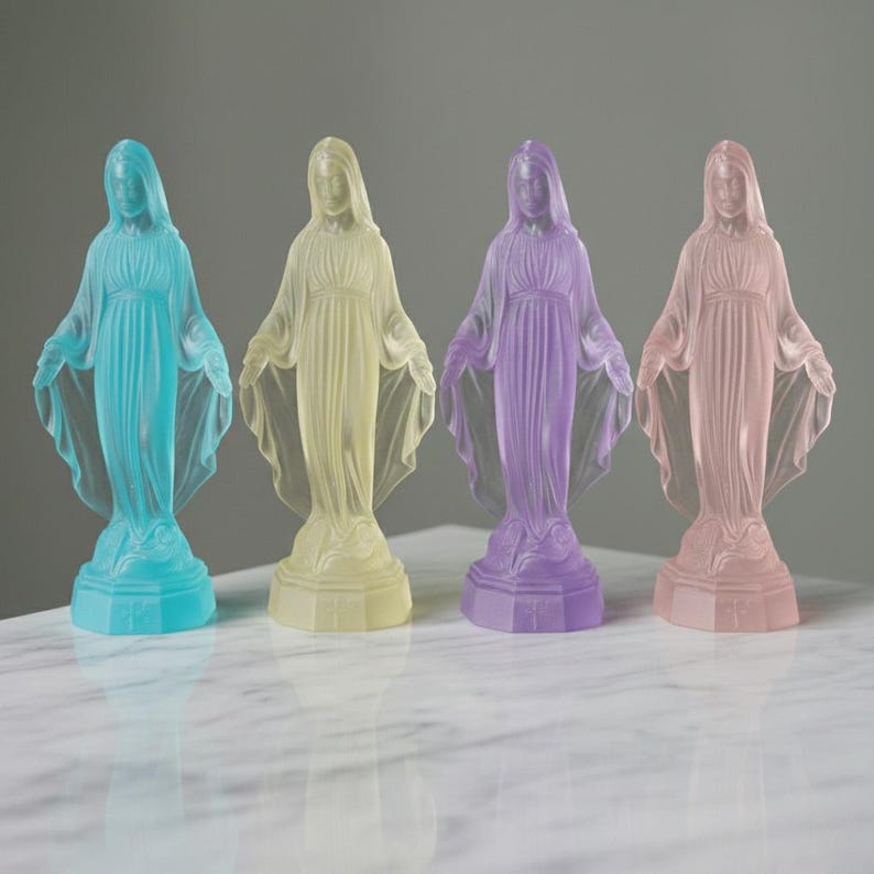 May include: Four translucent statues of the Virgin Mary standing in a row, each featuring a different pastel color: cyan, pale yellow, lavender, and soft pink. Each figure is depicted with arms outstretched, wearing a draped robe on a base.