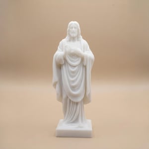 May include: A white marble statue of Jesus Christ, standing on a square base. The figure is depicted with long hair and a beard, wearing flowing robes. The hands are positioned near the chest. The statue is set against a neutral background.