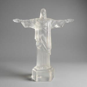 May include: A translucent statue of Christ the Redeemer with outstretched arms. The sculpture is mounted on a hexagonal base and stands on a gray background. The statue's robes and form are detailed, capturing the iconic pose.