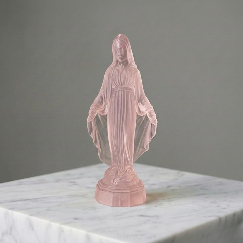 May include: A translucent pale pink glass figurine depicting a standing figure with arms outstretched, wearing a long, flowing robe with pleated details. The figure is positioned on a hexagonal base and features a smooth, frosted finish throughout.