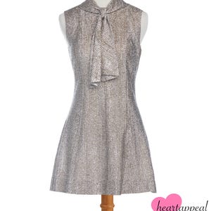 Puede incluir: Vestido plateado metálico sin mangas con cuello de lazo. El vestido tiene una falda acampanada y se exhibe en un maniquí. La tela tiene una textura acanalada vertical. Vestido de estilo vintage.