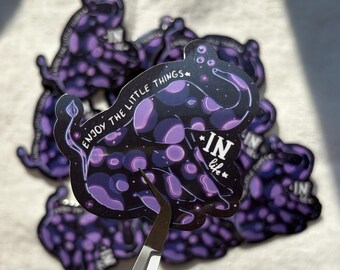 May include: A collection of stickers with a dark purple and black design. The stickers feature a stylized illustration with the text "ENJOY THE LITTLE THINGS" and "IN life". The stickers are held by a pair of silver tweezers.