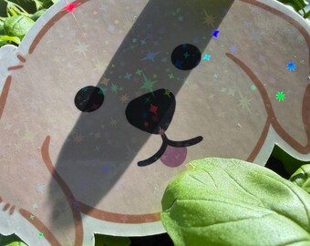 May include: A sticker featuring a cartoon dog face with black eyes, a black nose, and a pink tongue. The sticker has a holographic, glittery finish with star-shaped accents. The sticker is partially obscured by green basil leaves.