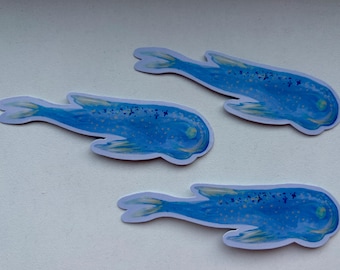 May include: Three blue whale stickers with a white border are arranged on a white surface. The whales have a watercolor effect with yellow and blue details. The stickers are a whimsical design.