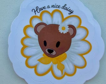 May include: A white, daisy-shaped sticker featuring a brown bear with a yellow bow and a small daisy on its head. The bear is centered within a yellow and white daisy design, with the phrase "Have a nice daisy" in cursive.